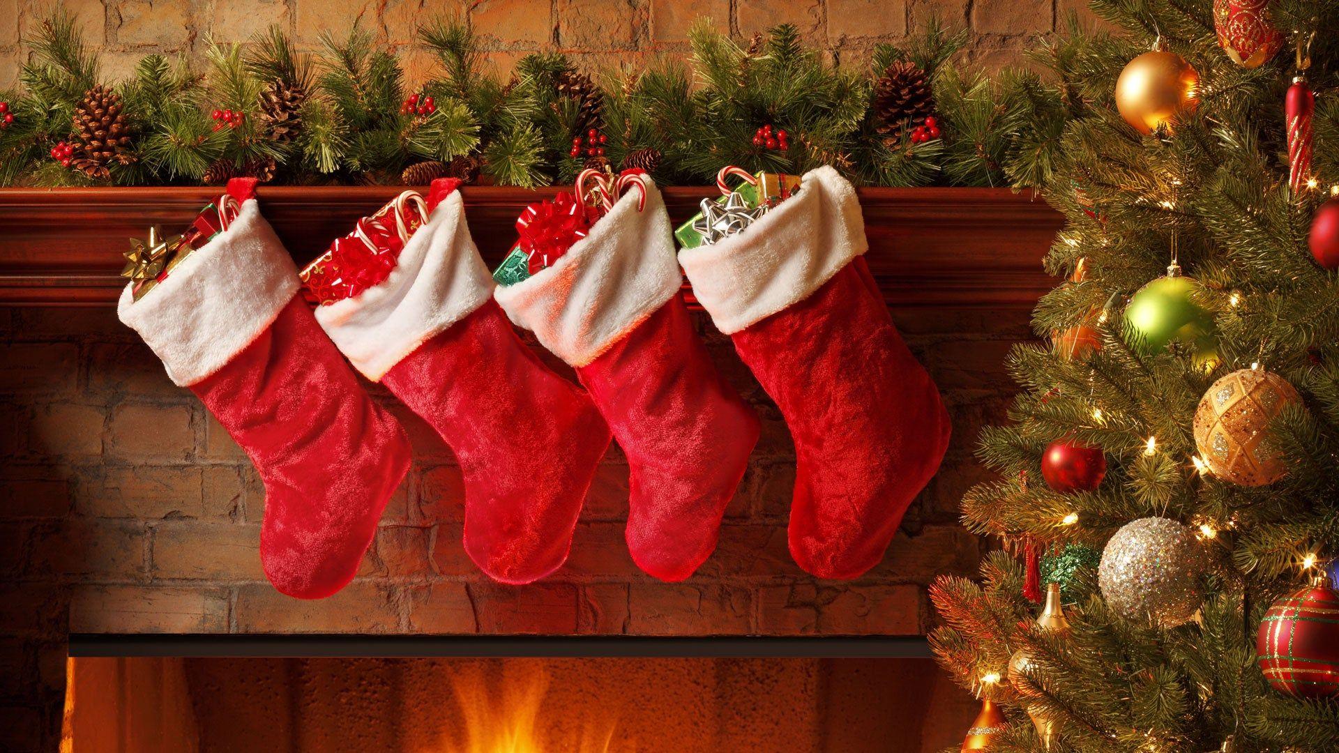 Four red stockings filled with gifts hanging above a fireplace next to a decorated Christmas tree.